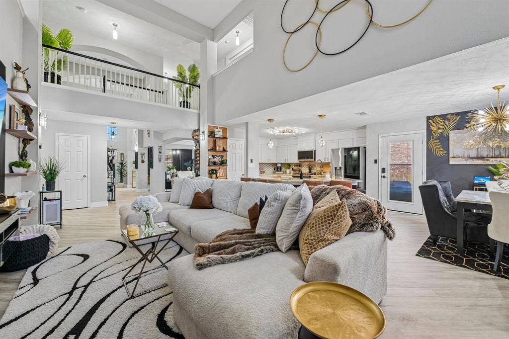 Open-concept living area featuring a double-height ceiling, upper-level balcony with white balustrade, wood-finish flooring, and a dining area with a gold chandelier
