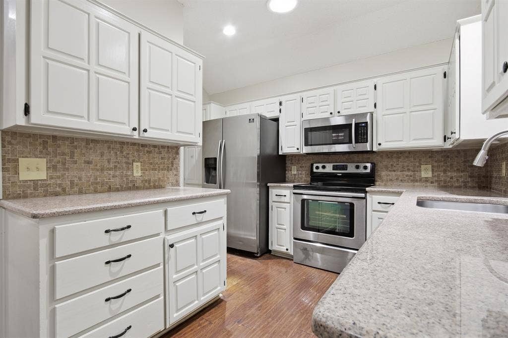 White paneled cabinetry, stainless steel appliances, speckled countertops, mosaic tile backsplash, and wood-finish flooring