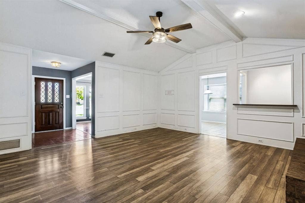 Vaulted ceiling with exposed beam, wood-finish flooring, decorative wall paneling, built-in display shelf, and an entry door with decorative glass inserts