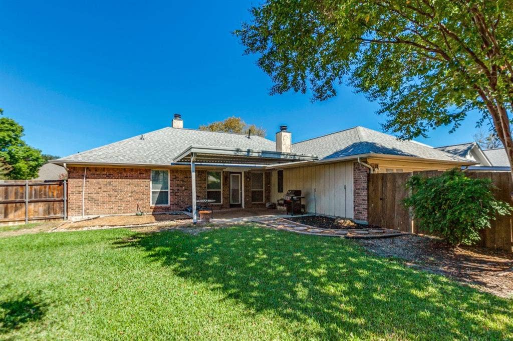 Expansive backyard featuring a manicured lawn, brick exterior, covered patio, privacy fencing, and mature trees