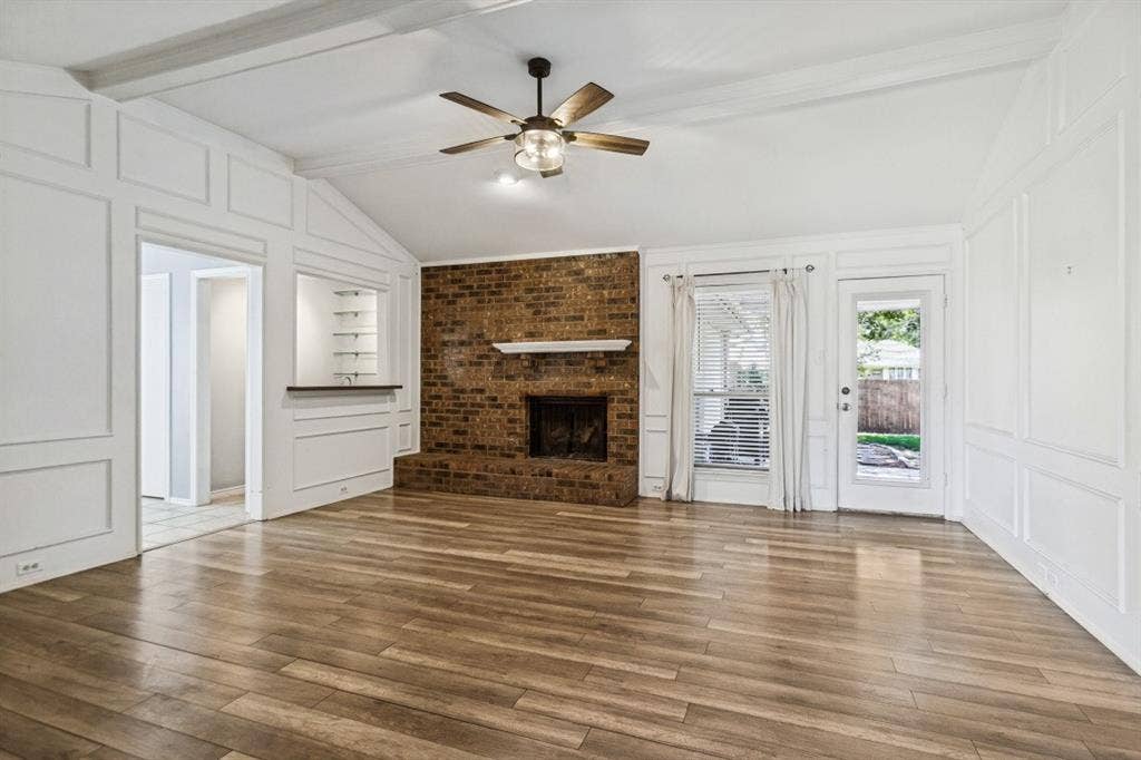 Vaulted ceiling with exposed beams, wood-finish flooring, brick fireplace with a white mantel, built-in shelving with recessed lighting, and a ceiling fan with integrated lighting