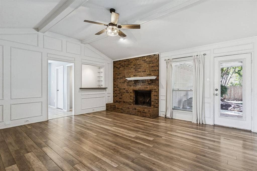 Spacious open-concept living area showcasing gleaming wood-finish flooring, a brick fireplace with integrated hearth, exposed ceiling beams, and abundant natural light from French doors and transom windows