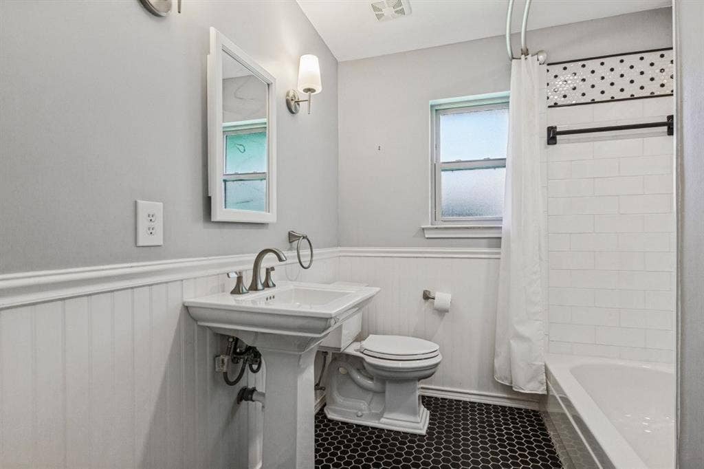 Pedestal sink with chrome fixtures, white wainscoting, black hexagonal floor tiles, subway tile shower surround, and a decorative tile accent