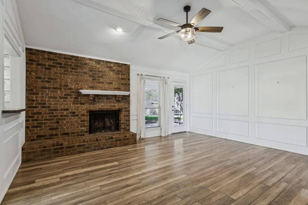 Spacious open-concept living area featuring a striking exposed brick fireplace with mantel, warm wood-finish flooring throughout, recessed lighting, ceiling fan with integrated light fixture, and glass-paneled French doors providing natural light and access to outdoor space