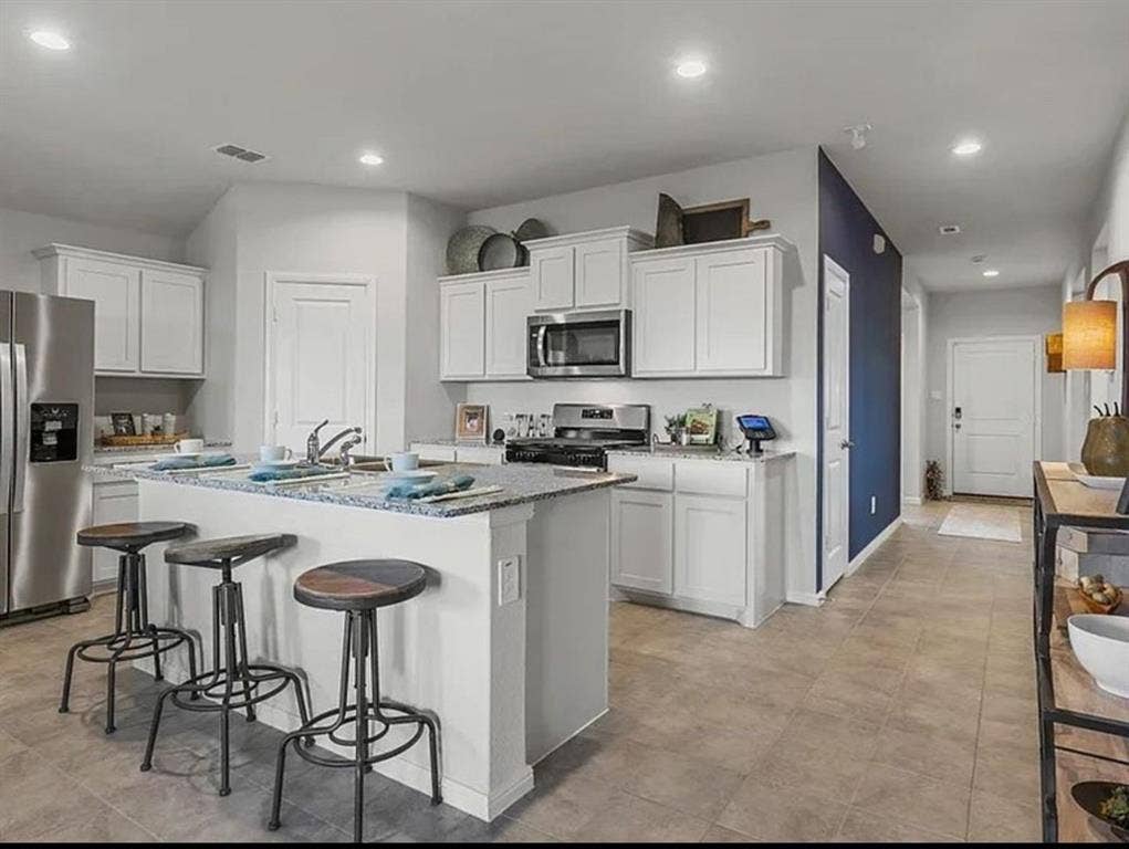 Staged Kitchen with a kitchen bar, stainless steel appliances, recessed lighting, white cabinets, and a kitchen island with sink. Hallway wall is not blue.