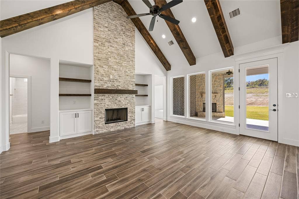 104 Oak Meadow Lane, Weatherford, TX 76085 Vaulted ceilings with exposed wood beams, a floor-to-ceiling stone fireplace, built-in cabinetry with wood-finish shelving, wood-finish tile flooring, and multiple large windows with exterior access