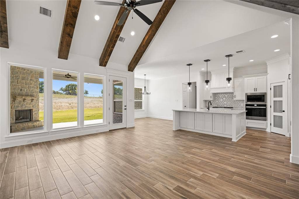 104 Oak Meadow Lane, Weatherford, TX 76085 Vaulted ceiling with exposed wood beams, stone fireplace, wood-finish flooring, white cabinetry, and a kitchen island with pendant lighting