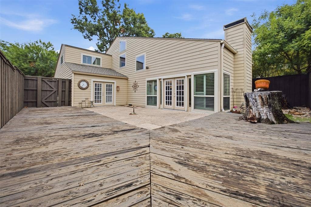 Expansive composite deck with wood-finish flooring overlooking the rear grounds, anchored by a contemporary home featuring cream-toned siding, multiple sets of French doors with operable shutters, and mature landscaping with established trees providing natural screening and privacy