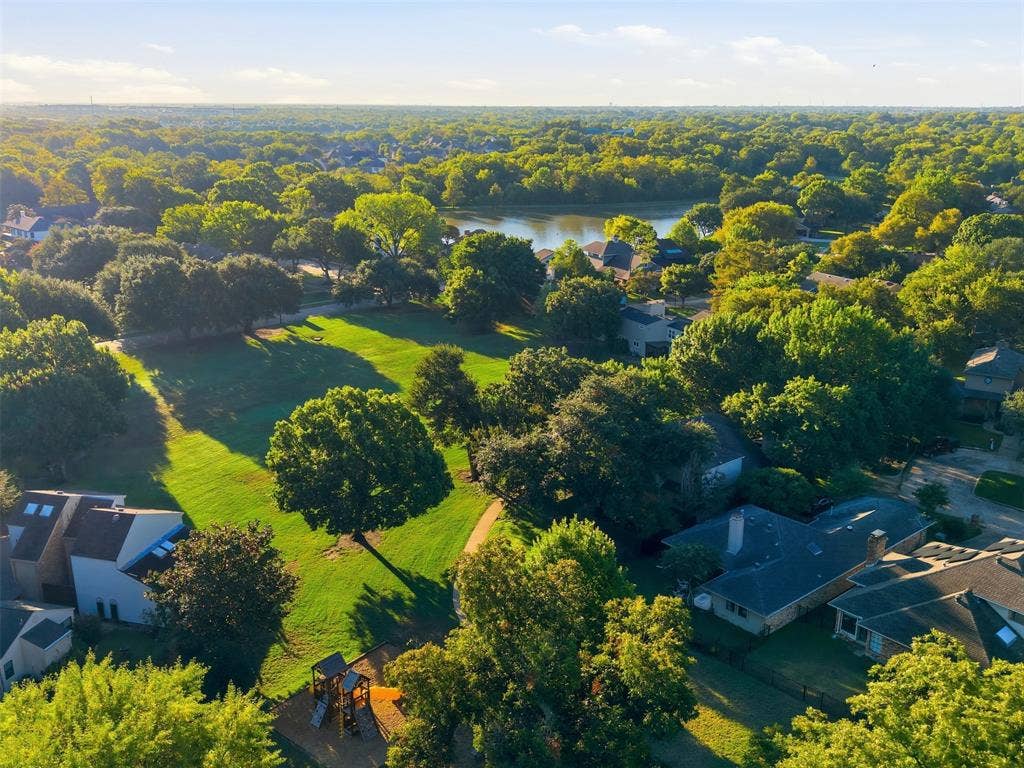 Expansive green space, mature trees, playground equipment, paved walking path, and a distant lake