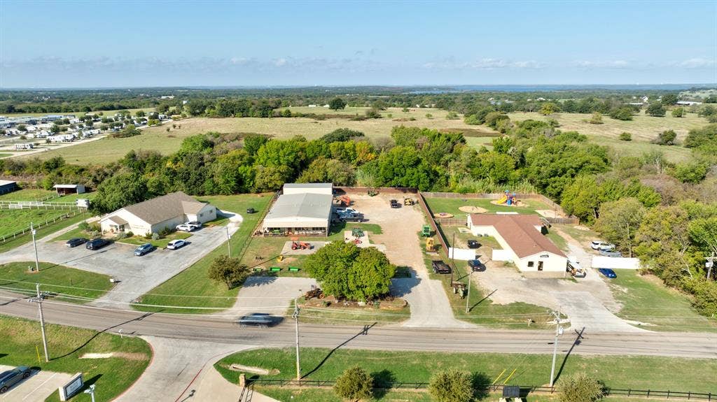 Aerial perspective of a multi-structure property featuring paved parking surfaces, a large metal-framed industrial building, a primary building with a shingled roof, and a secondary structure with a fenced perimeter