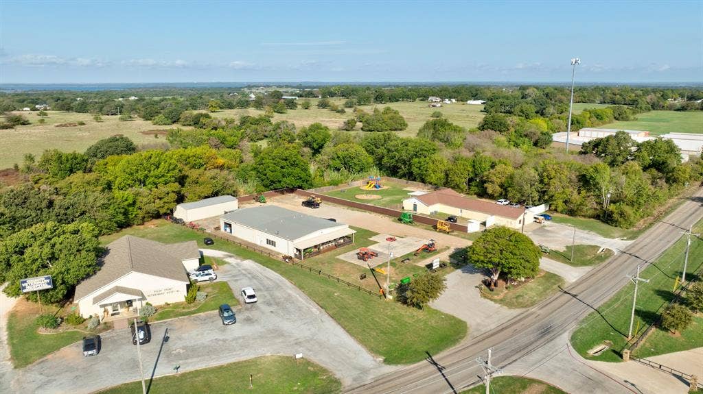 Aerial perspective of a multi-structure property featuring paved parking areas, metal-sided outbuildings, and open acreage with mature perimeter trees