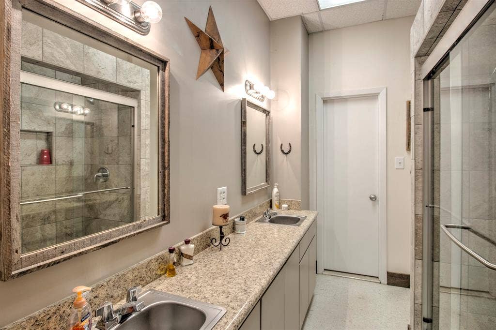Extended vanity with stone-composite countertop, dual undermount stainless steel sinks, glass-enclosed shower with tiled surround, and rustic wood-trimmed wall mirrors