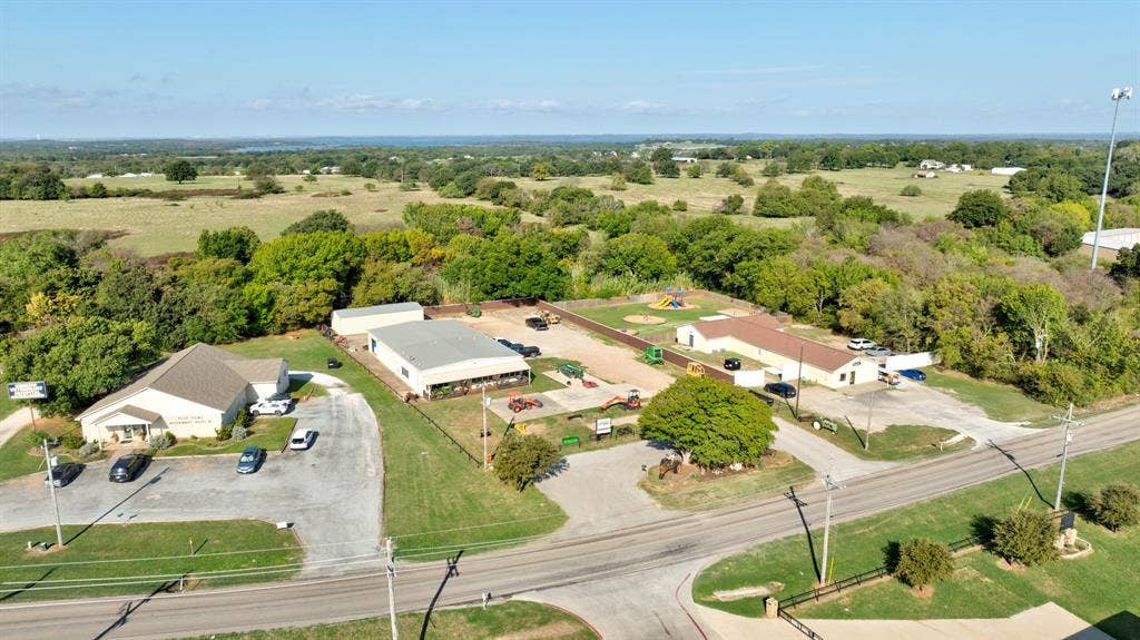 Aerial perspective of a multi-structure property featuring paved parking areas, metal-roofed buildings, a fenced perimeter, and mature landscaping