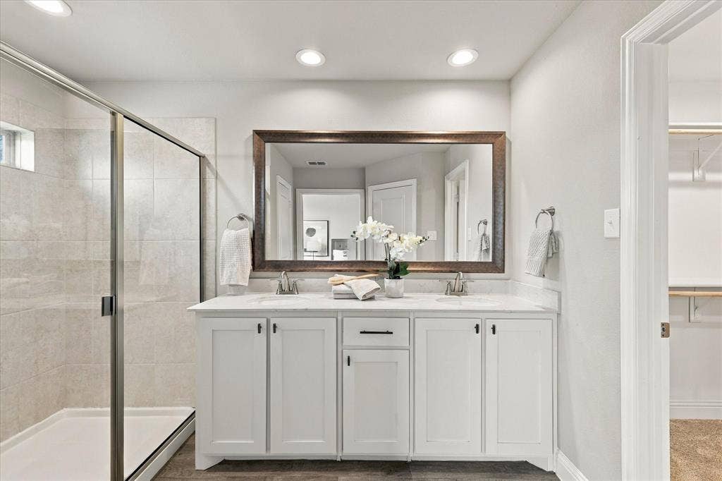 Dual vanity with white cabinetry and integrated sinks, large framed mirror, glass-enclosed shower with tiled walls, recessed lighting, and wood-finish flooring