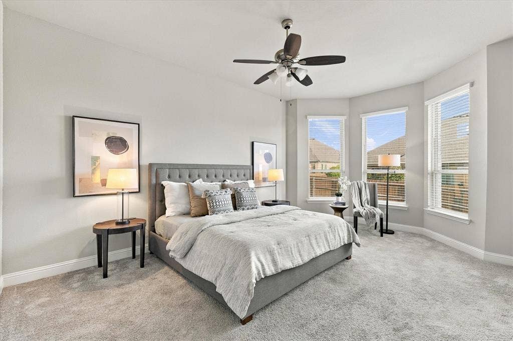 Bay window feature with three large windows, light gray walls, light gray carpet flooring, ceiling fan with integrated lighting, and a tufted upholstered bed frame