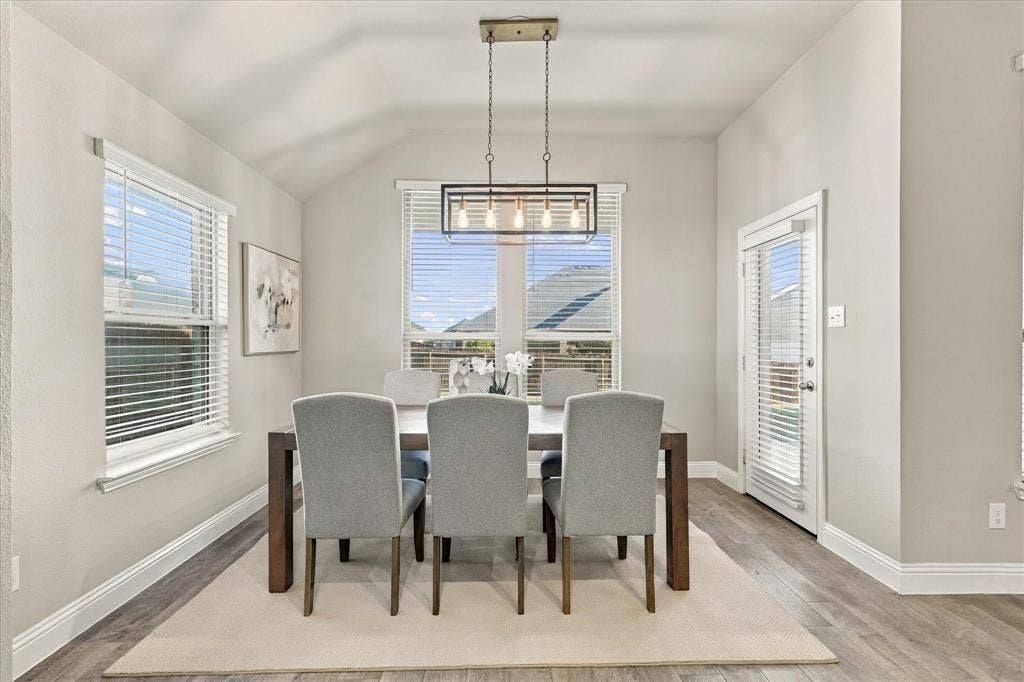 Vaulted ceiling, wood-finish flooring, two windows with blinds, a glass-paneled door, and a rectangular hanging light fixture