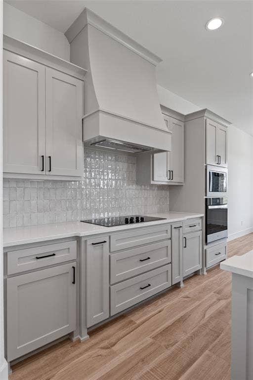 Light gray cabinetry with contemporary black hardware, white solid surface countertops, a mosaic tile backsplash, built-in cooktop, and wood-finish flooring