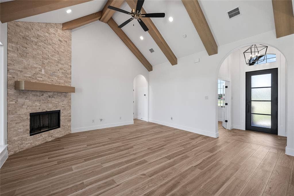 Vaulted ceiling with exposed wood beams, stone-clad fireplace with mantel, wood-finish flooring, recessed lighting, and an arched entry with a glass-paneled door