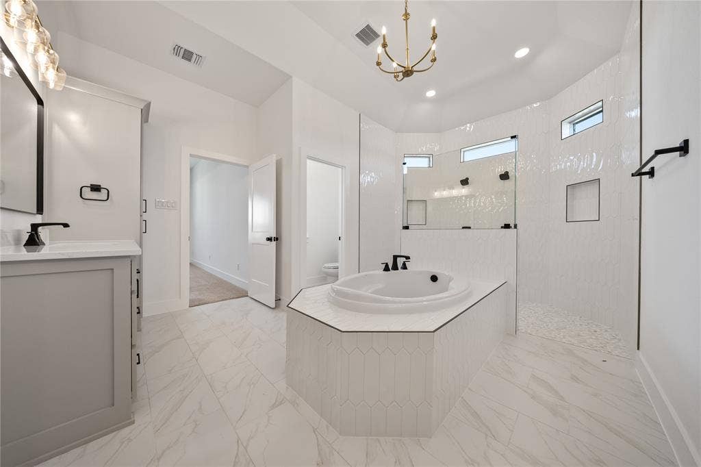 White tile flooring, built-in soaking tub, walk-in shower with glass enclosure, single vanity with white countertop, and brass chandelier