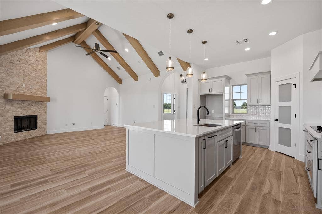 Vaulted ceilings with exposed wood beams, stone fireplace with a wood mantle, wood-finish flooring, kitchen island with a built-in sink and pendant lighting, and light grey cabinetry