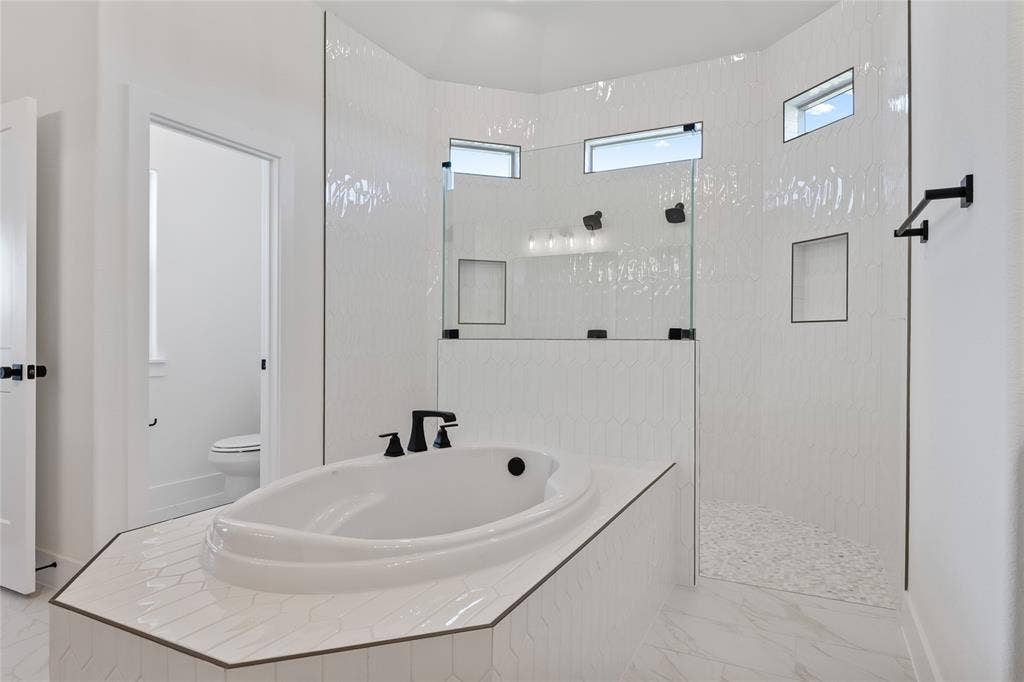Soaking tub with matte black fixtures, an expansive walk-in shower with glass enclosure, white hexagon tile walls, and large format floor tile