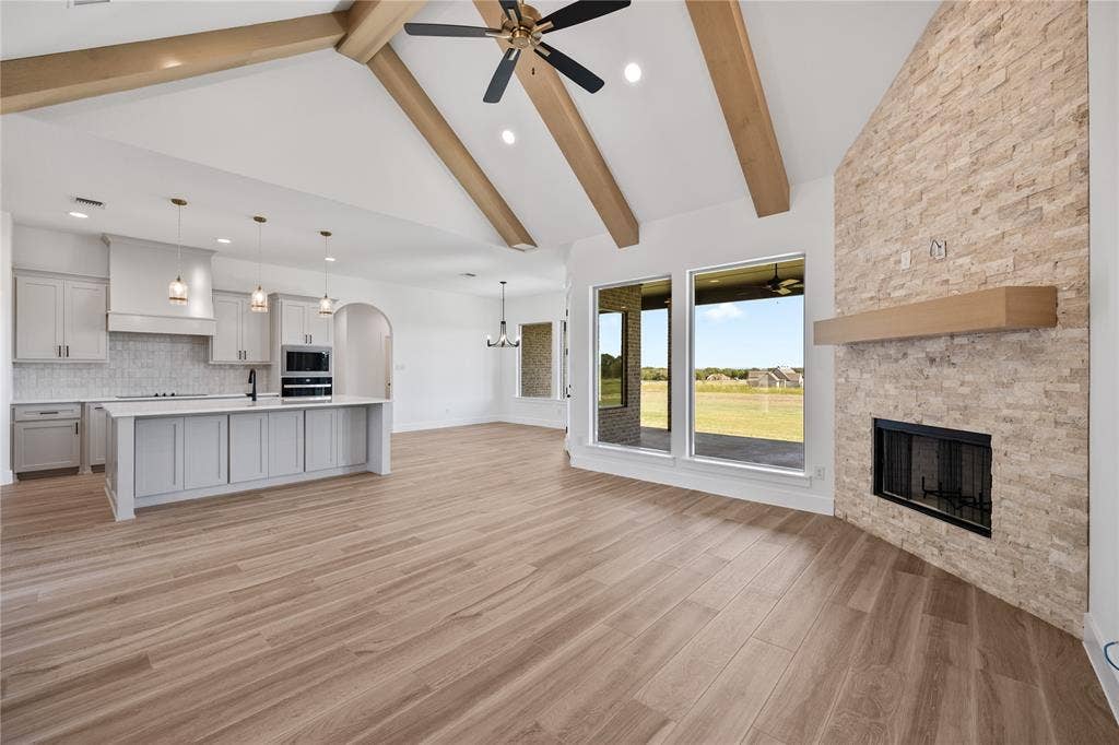 Vaulted ceiling with exposed wood beams, wood-finish flooring, stone-clad fireplace with wood mantel, large windows, and a kitchen island with pendant lighting