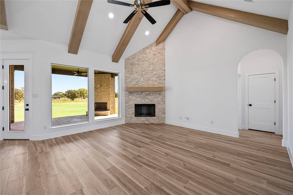 Vaulted ceiling with exposed wood beams, stone-faced fireplace with wood mantel, large fixed-pane windows, wood-finish flooring, and an arched entryway