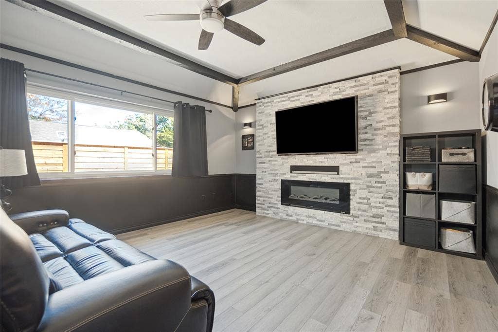 7414 Windmill Lane, Garland, TX 75044 Light wood-finish flooring, accent wall with stacked stone-look tile, built-in electric fireplace, recessed wall lighting, and exposed ceiling beams