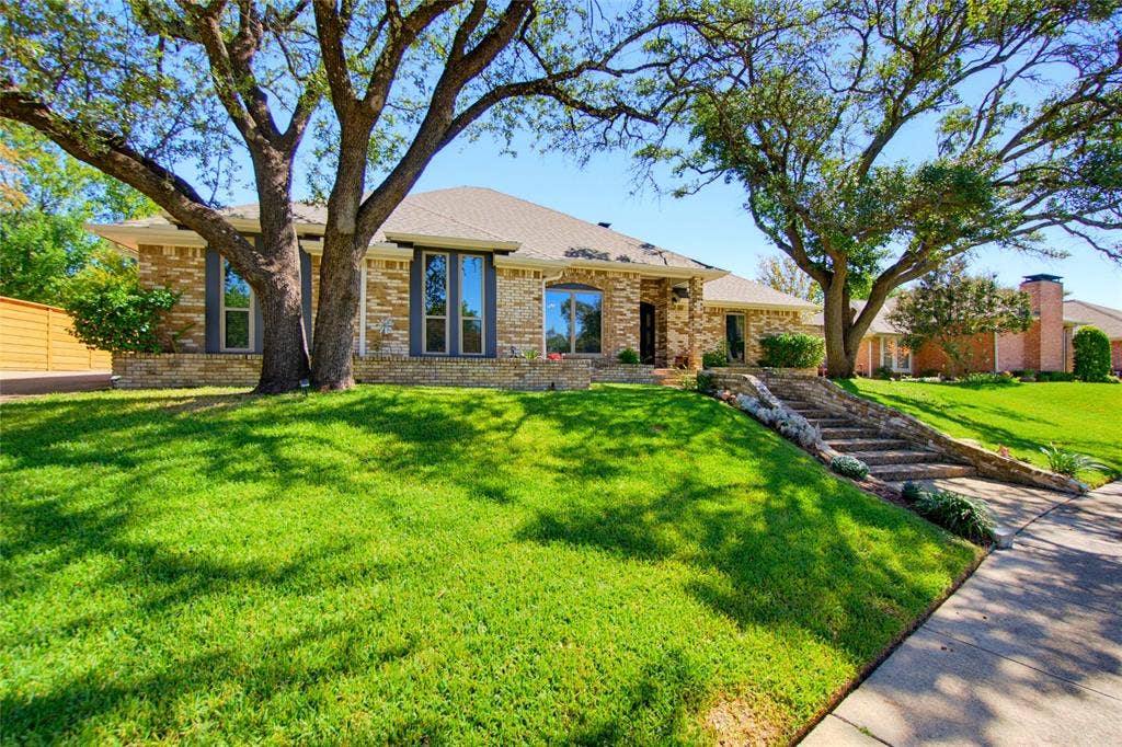7414 Windmill Lane, Garland, TX 75044 Brick and stone facade residence featuring a sloped front lawn, mature trees, and a stone-lined staircase