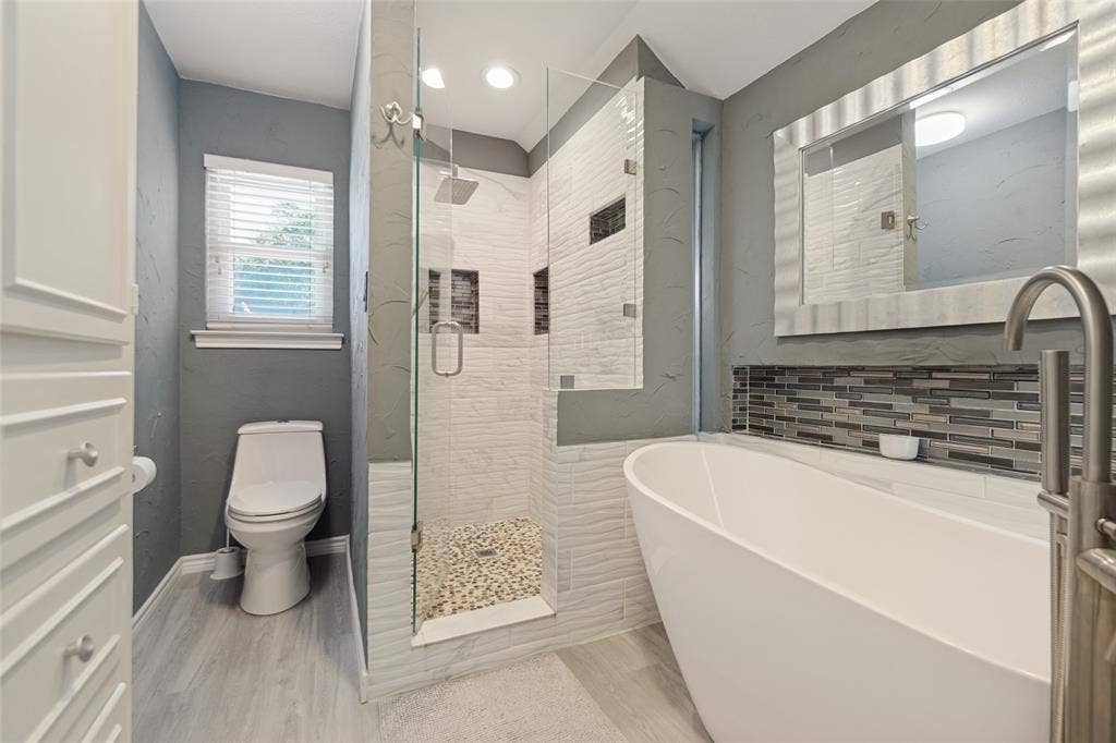 7414 Windmill Lane, Garland, TX 75044 Freestanding soaking tub, glass-enclosed shower with pebble floor, rain showerhead, mosaic tile accent, and light wood-finish flooring