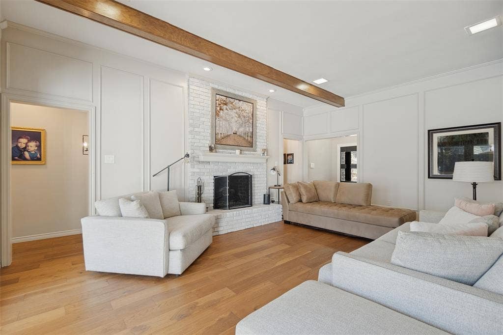 7414 Windmill Lane, Garland, TX 75044 Spacious living area featuring wood-finish flooring, a white brick fireplace with a hearth, recessed lighting, decorative wall paneling, and an exposed ceiling beam
