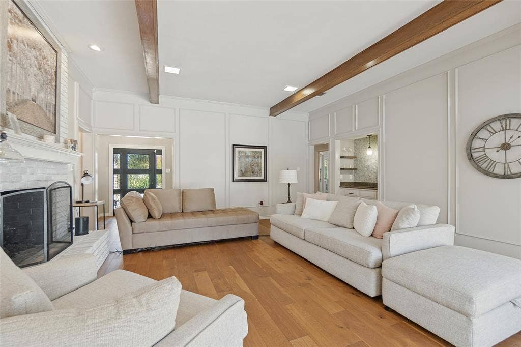 7414 Windmill Lane, Garland, TX 75044 Wood-finish flooring, white paneled walls, white brick fireplace with a black screen, exposed wood ceiling beams, and a built-in recessed bar area with shelving
