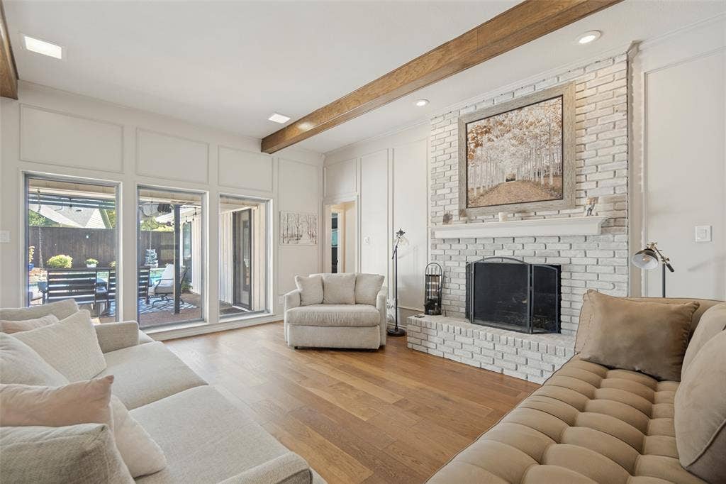 7414 Windmill Lane, Garland, TX 75044 Hardwood flooring throughout, a white brick fireplace with a mantel, exposed wood beams, and a wall of glass doors opening to the exterior