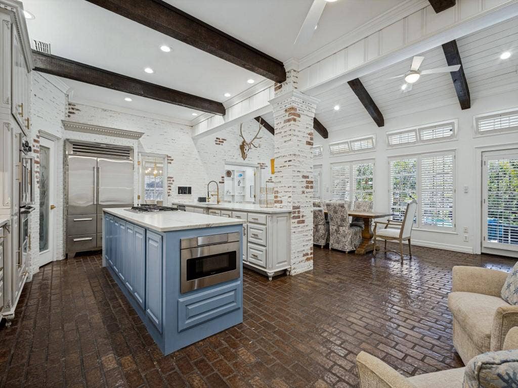 Spacious kitchen featuring exposed wood beams, brick accent walls, a kitchen island with built-in microwave, stainless steel appliances, and brick flooring