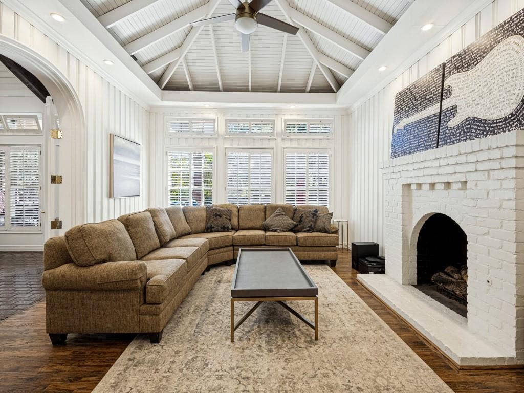 Vaulted ceiling with exposed beams, wood-finish flooring, white painted brick fireplace, recessed lighting, and wall paneling