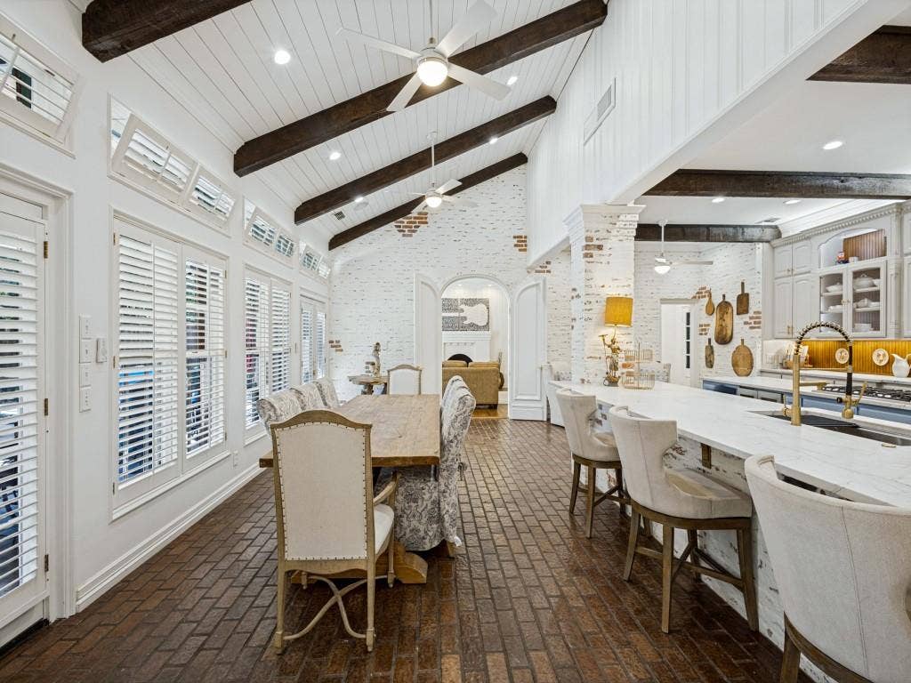 Vaulted ceiling with exposed wood beams, brick-finish flooring, plantation shutters, painted brick wall finish, and a kitchen island with stone countertop