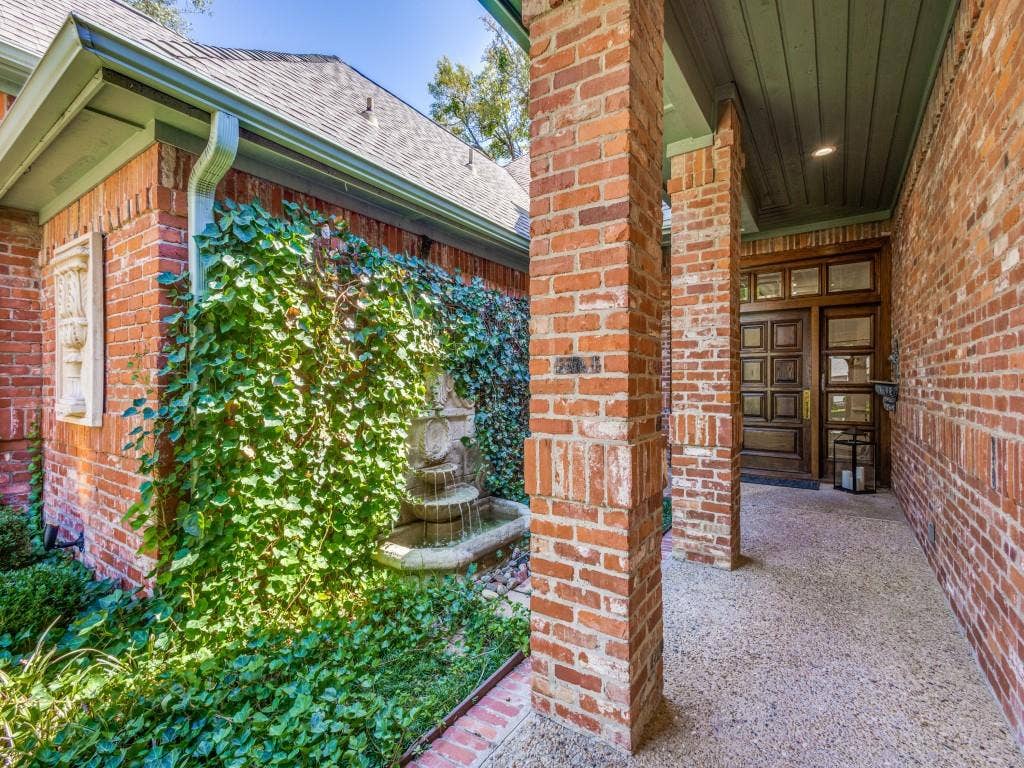 Brick exterior featuring a multi-paneled wood entry door, covered porch with exposed aggregate flooring, and a decorative wall fountain with lush greenery