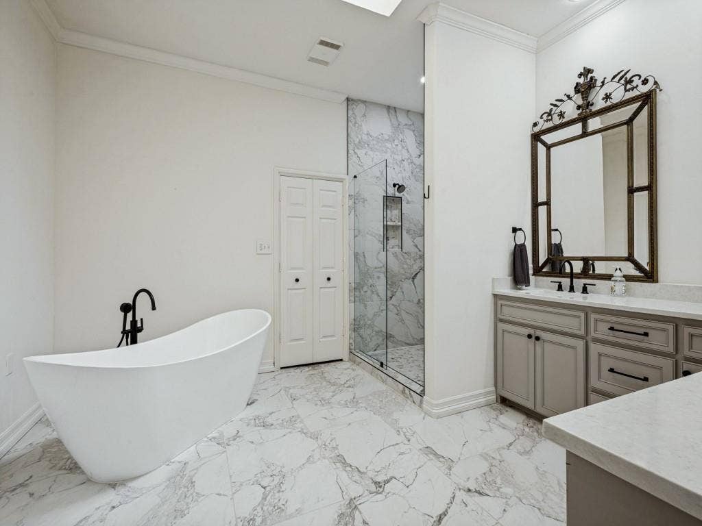 Freestanding soaking tub with a matte black faucet, glass-enclosed shower with tile surround, double vanity with light gray cabinetry, light-toned countertops, and marble-look tile flooring