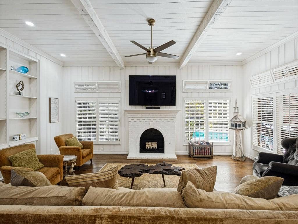 Living area featuring a white brick fireplace, wood-finish flooring, built-in shelving, wood-beam ceiling, and multiple windows with white louvered blinds