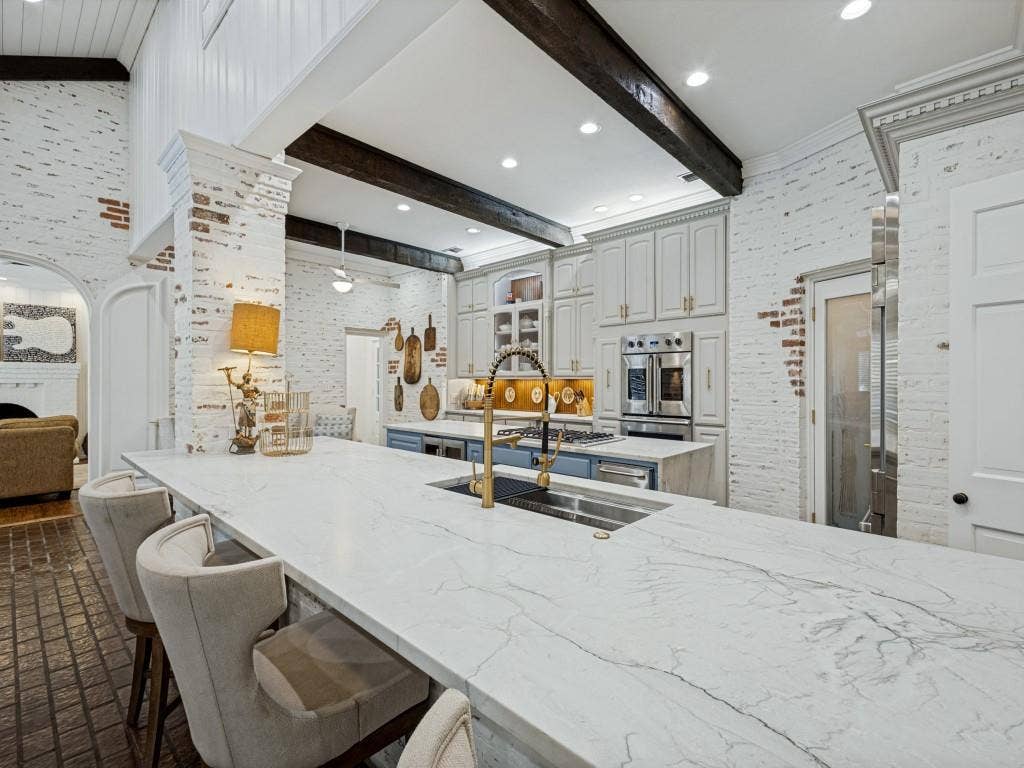 Expansive kitchen island with a stone-finish countertop, white-washed brick walls, vaulted ceiling with exposed wood beams, built-in stainless steel double ovens, and light-colored cabinetry
