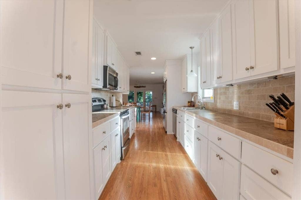 Galley kitchen featuring white cabinetry, wood-finish flooring, stainless steel appliances, a tile backsplash, and light-colored countertops