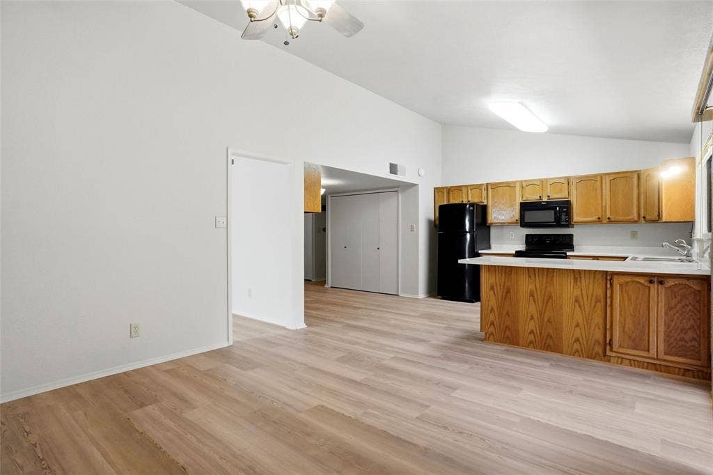 Vaulted ceiling with ceiling fan, wood-finish flooring, kitchen with light wood cabinetry, white countertops, and black appliances