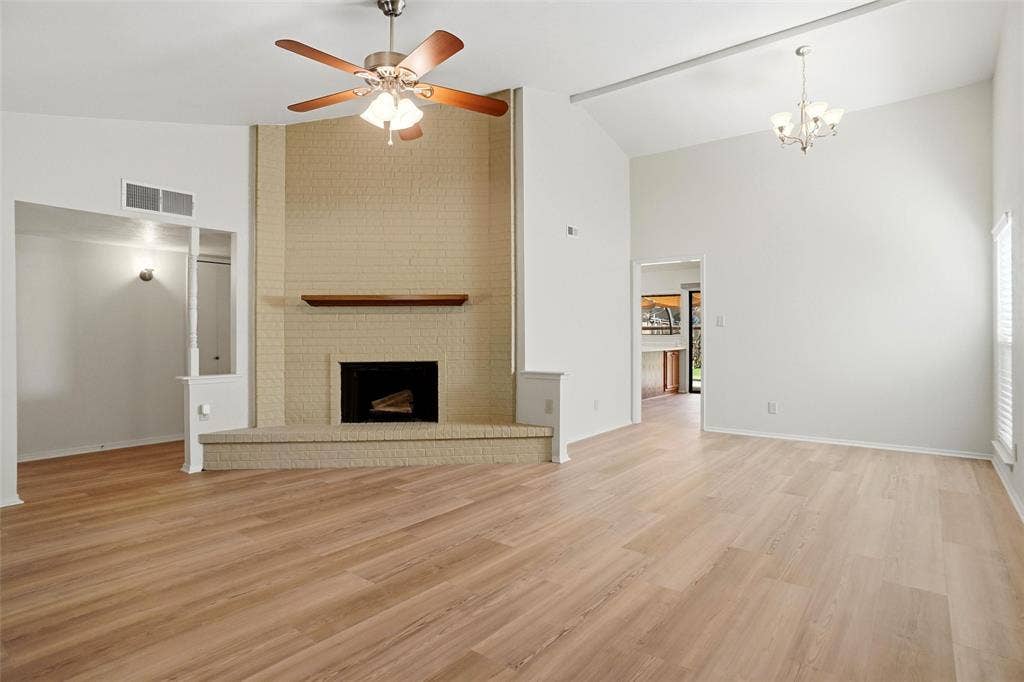Vaulted ceiling with exposed beam and chandelier, wood-finish flooring, brick fireplace with wood mantel, ceiling fan with integrated lighting, and window with blinds