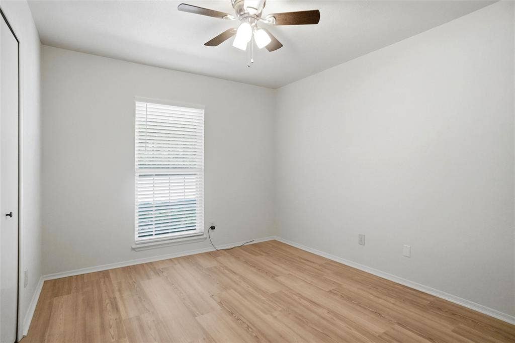 Hardwood-finish flooring, single window with blinds, ceiling fan with light fixture, white walls, and a white door with a black handle