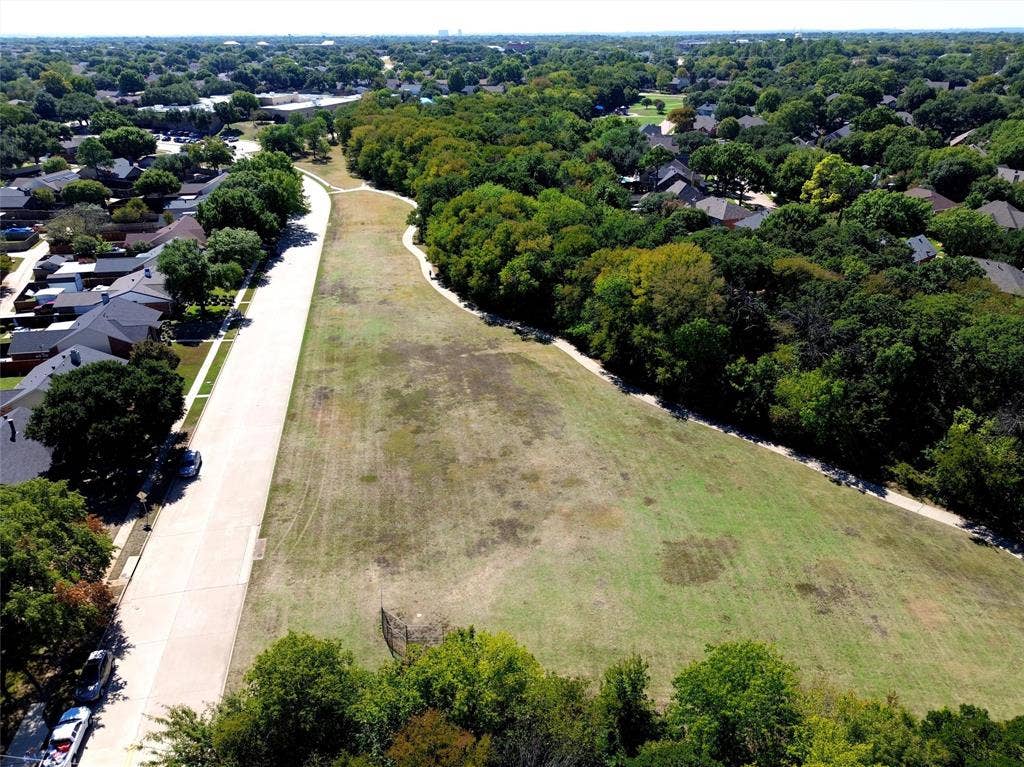 Expansive green space bordered by mature trees and residential properties, featuring a paved roadway and a winding pathway