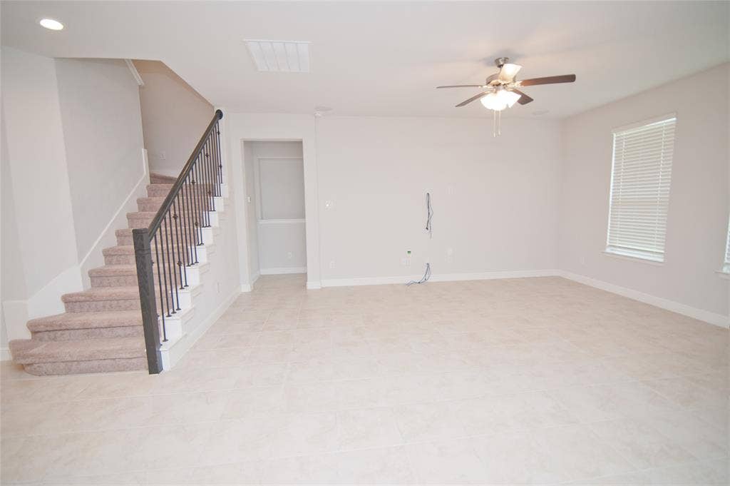 1021 Gillespie Drive, Fort Worth, TX 76247 Spacious room featuring tile flooring, a staircase with carpeted treads and wrought iron balusters, a ceiling fan with integrated lighting, and multiple windows with blinds