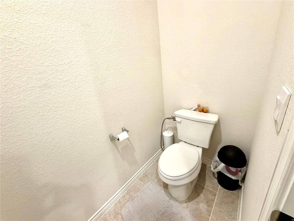 White commode with a bidet attachment, light-toned textured walls, and tiled flooring