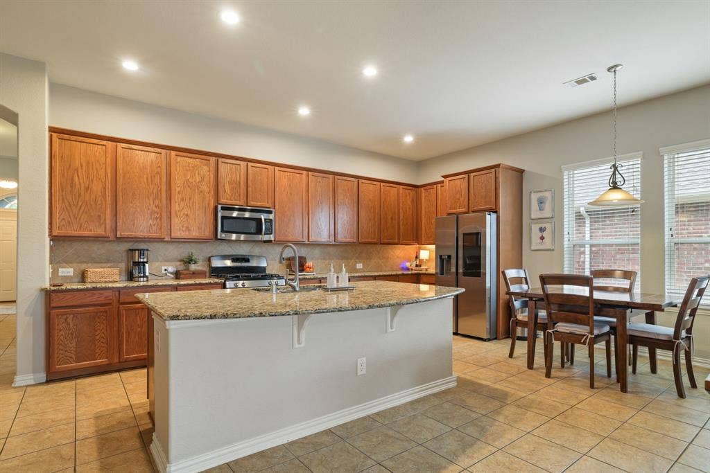 1213 Egret Court, Little Elm, TX 75068 Kitchen featuring wood cabinetry, stainless steel appliances, a center island with a granite-style countertop, and tile flooring