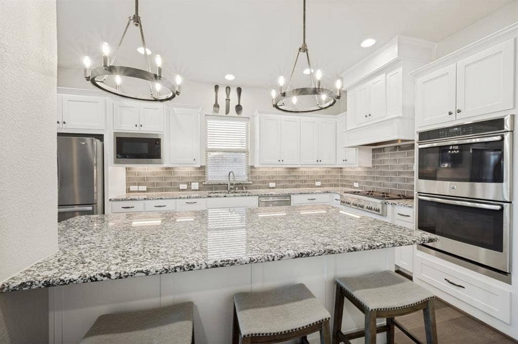 2721 Old Shire Path Road, Prosper, TX 75078 White cabinetry, stone countertops, stainless steel appliances, tile backsplash, and two chandelier light fixtures