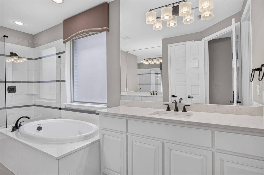 702 Callaway Drive, Allen, TX 75013 Built-in soaking tub with surround tiling, glass-enclosed shower, white vanity with stone countertop, undermount sink, and dark bronze fixtures